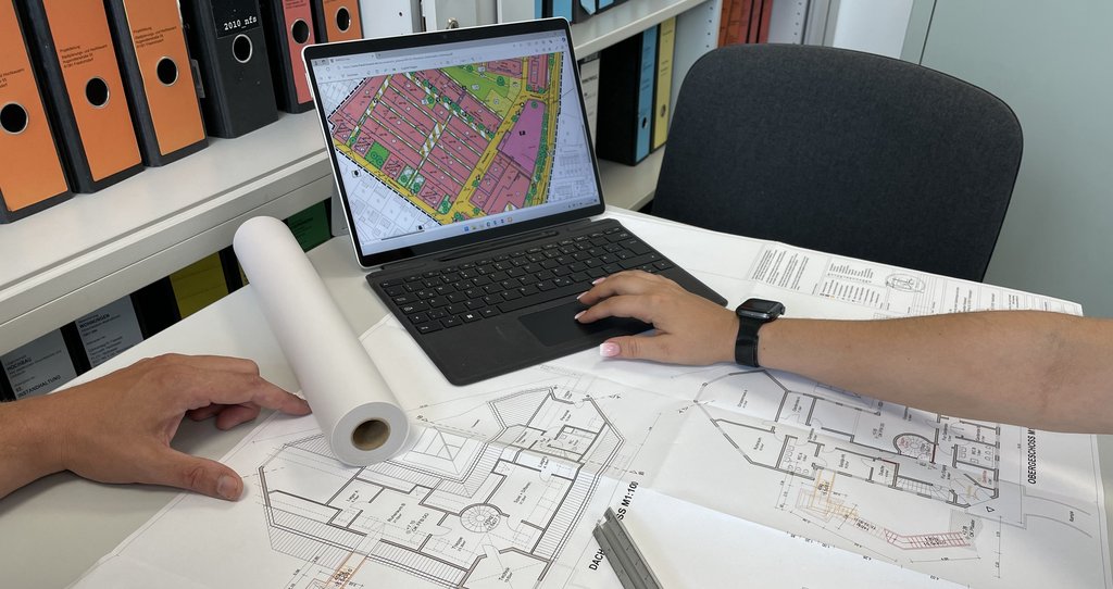 Auf dem Bild ist ein Bauplan auf einem Schreibtisch sowie ein Laptop zu sehen.
