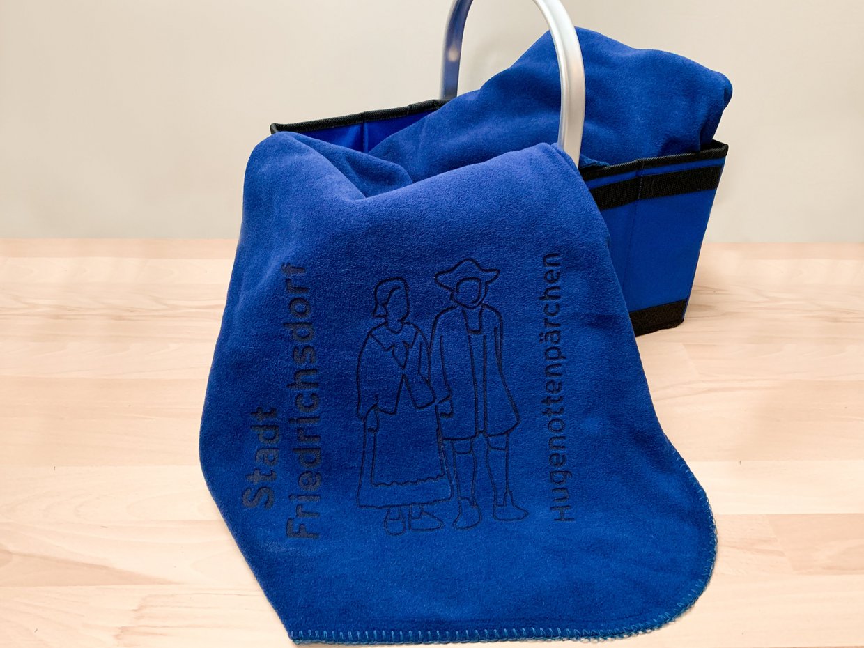 Auf der Darstellung ist eine blaue Fleecedecke zu sehen, die mit dem Hugenottenpärchen der Stadt Friedrichsdorf verziert ist.