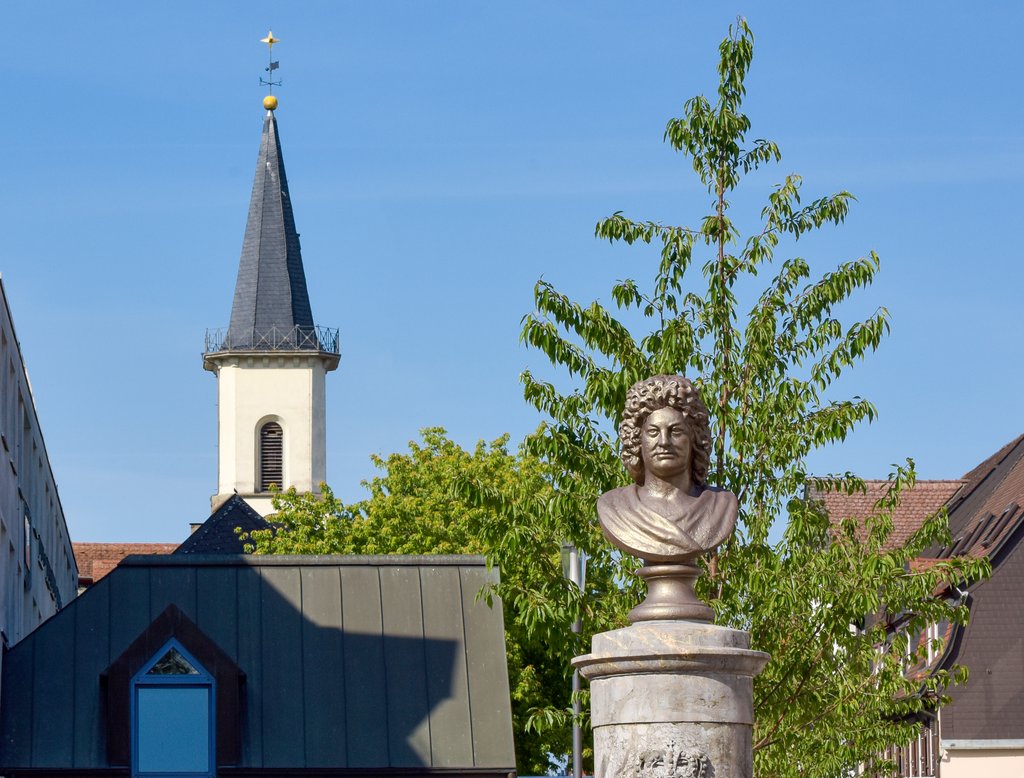 Auf der Darstellung sind die Büste des Landgrafen Friedrich II. von Hessen-Homburg und der Kirchturm der Evangelischen Kirche Friedrichsdorf zu sehen.