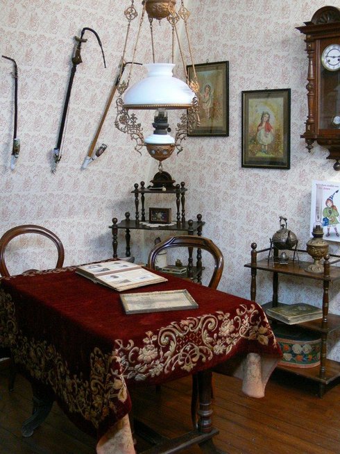 Ein Raum im Stil der Gründerzeit mit rot gepolsterten Stühlen, einem Tisch mit rotem Tuch und Fransen, darauf ein üppig verziertes Kaffeeservice zur Goldhochzeit. Eine Puppe sitzt in einem Kinderhochstuhl, an der Wand hängen eine Standuhr und alte Lampen. Der Raum wirkt warm, wohnlich und erinnert an eine gute Stube um 1900.