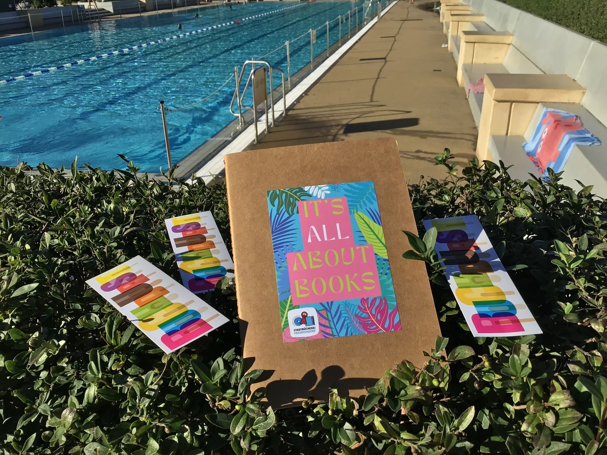 „Buchdurst“-Abschlussparty im Freibad Das Bild zeigt ein Schwimmbecken des Friedrichsdorfer Freibads und im Vordergrund Werbematerialien zum Sommerleseclub „Buchdurst“ der Stadtbücherei Friedrichsdorf.