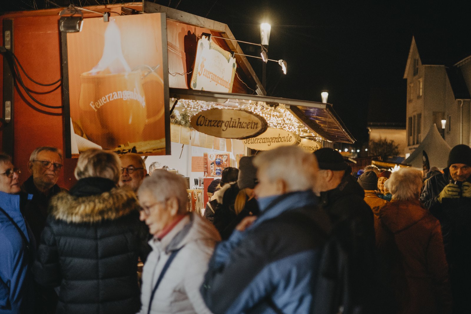 Weihnachtsmarkt 2024_Menschen_Feuerzangenbowle Das Bild zeigt einen Weihnachtsmarkt bei Abend. Es ist dunkel und im Vordergrund drängen sich Menschen um eine Feuerzangenbowle Bude.