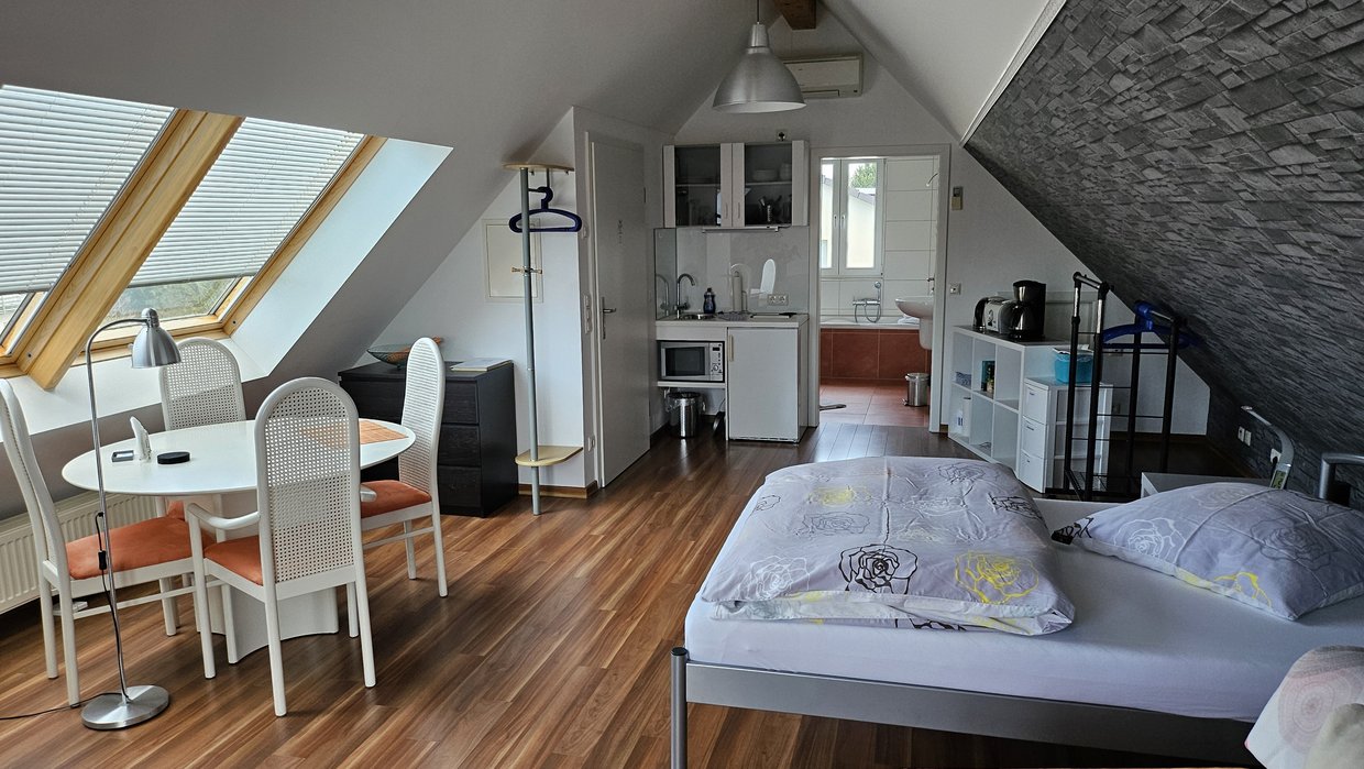 Auf der Darstellung ist eine Ferienwohnung im Dachgeschoss mit Doppelbett, Sitzplatz und Küchenzeile zu sehen.