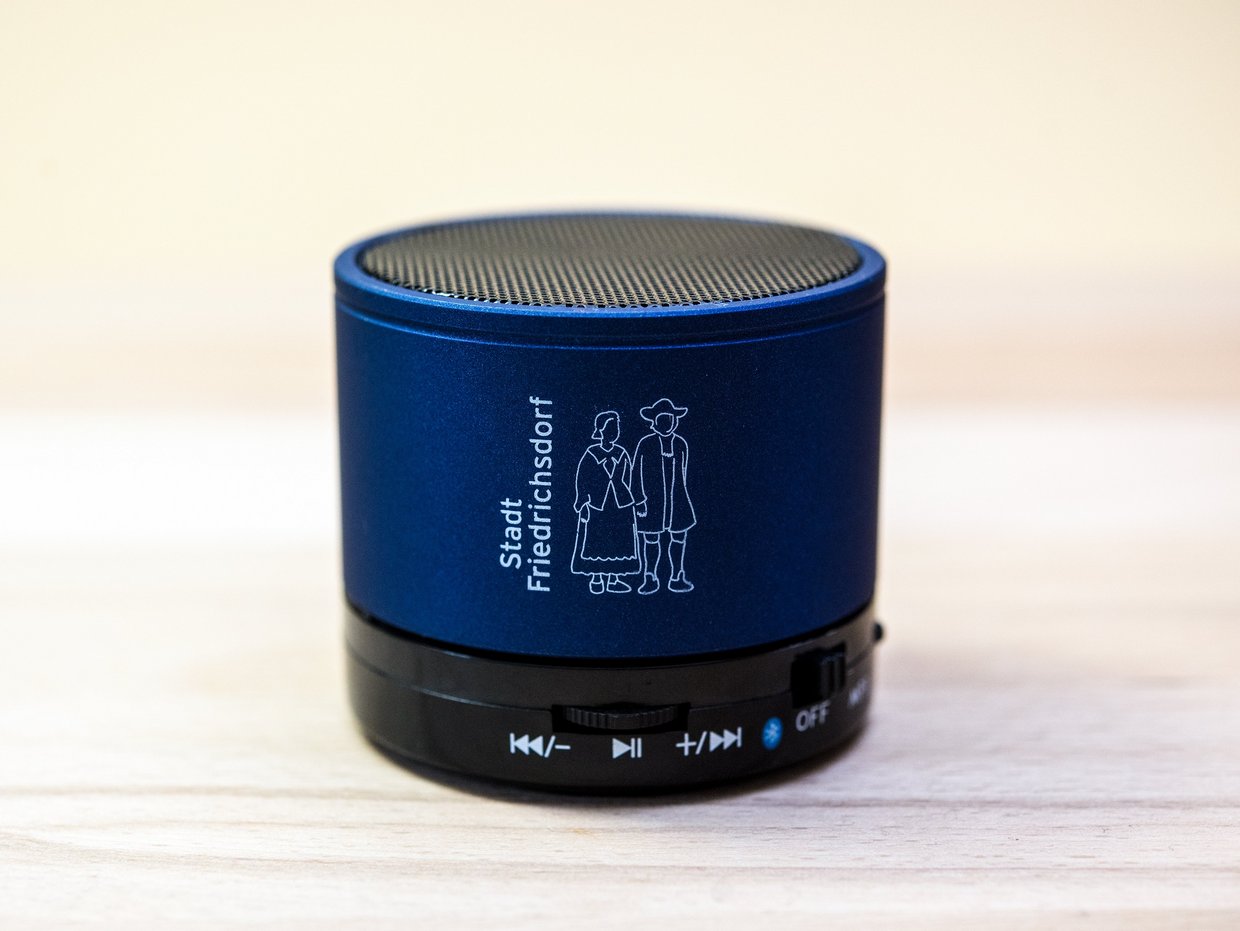 Auf der Darstellung ist ein blauer Bluetooth-Lautsprecher zu sehen, der mit dem Hugenottenpärchen in weiß verziert ist.