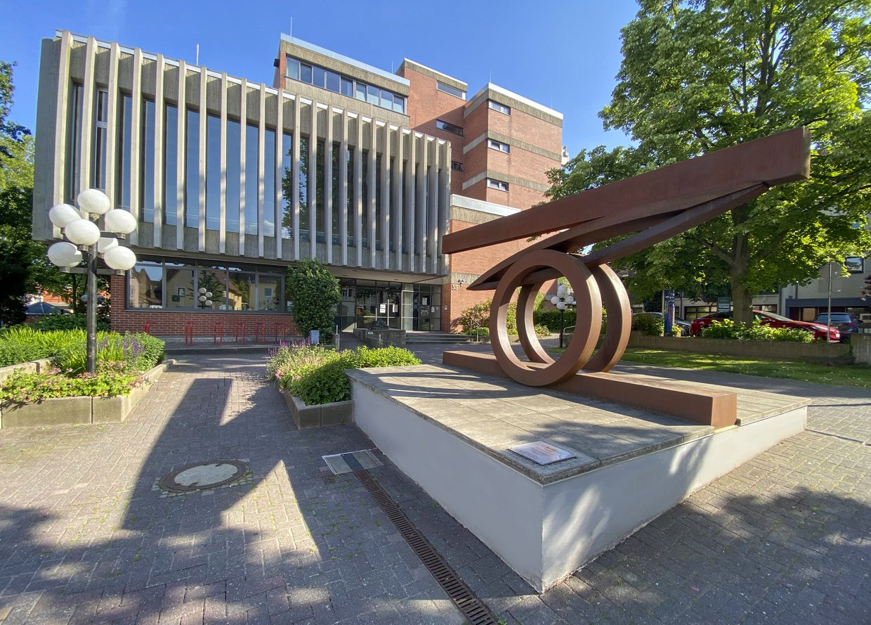 Auf der Darstellung ist das Rathaus Friedrichsdorf zu sehen, im Vordergrund die Skulptur „Im Gleichgewicht“ von Claus Bury.