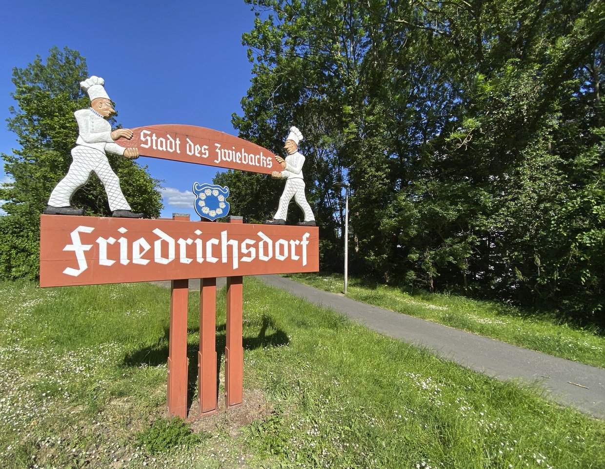 Auf der Darstellung ist das Holzschild „Stadt des Zwiebacks“ mit zwei Zwiebackbäckerfiguren zu sehen.