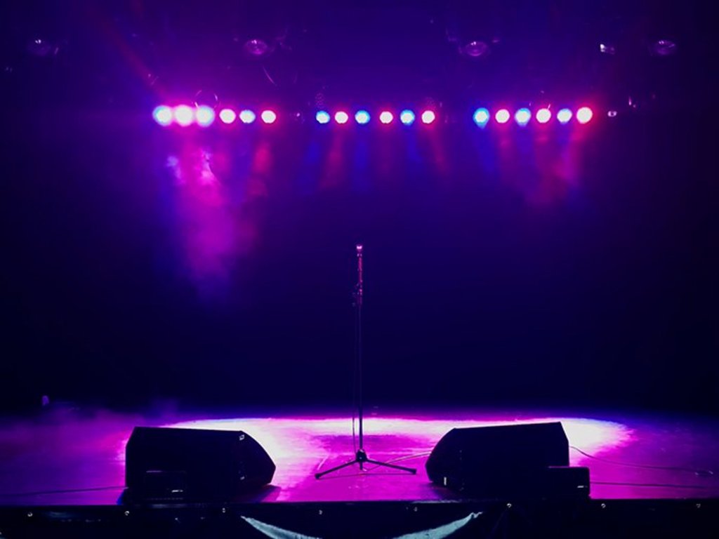 Auf der Darstellung ist die in pinkes und blaues Scheinwerferlicht getauchte Bühne im Forum Friedrichsdorf zu sehen, in der Mitte ein Mikrofonständer umrahmt von zwei Monitorboxen.