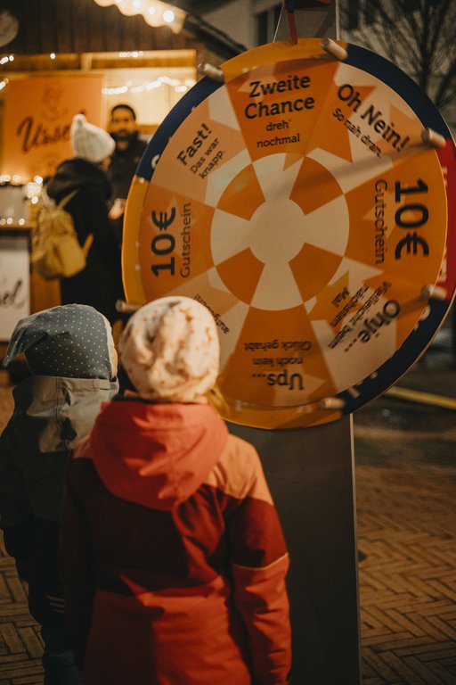 Kinder in Winterjacken stehen mit dem Rücken zum Betrachtenden vor Glücksrad.