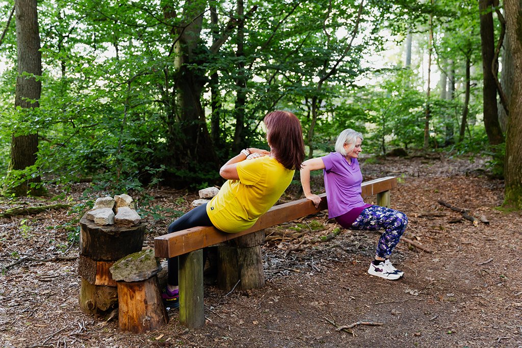 Auf der Darstellung sind zwei Personen zu sehen, die an einer Station des Trimm-Dich-Pfads Dehnübungen machen.