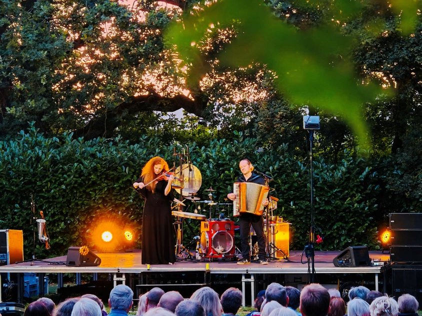 Eine Bühne im Freien mit Hecken und Bäumen im Hintergrund. Auf der Bühne stehen zwei Musiker.