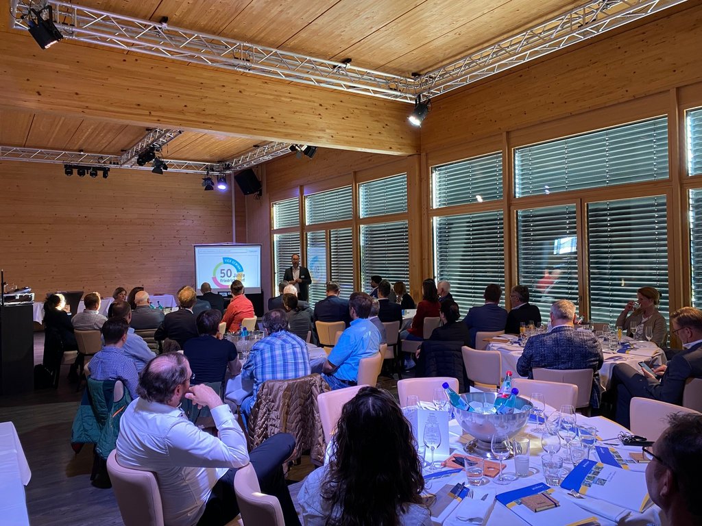 Mehrere Personen hören dem Bürgermeister bei einer Präsentation zum Standort Friedrichsdorf in einem festlichen Rahmen zu.