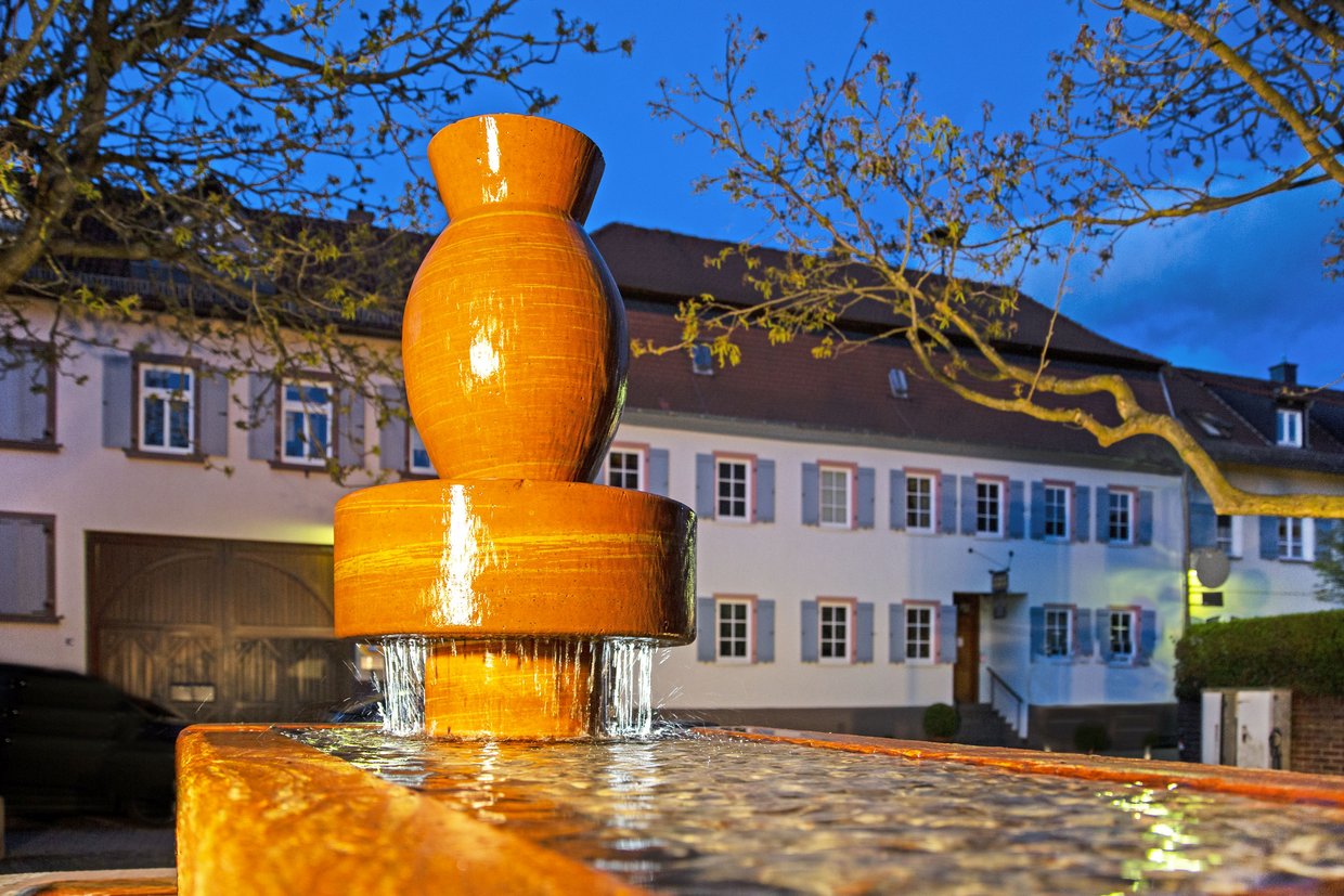 Auf der Darstellung ist das Heimatmuseum in Seulberg in der Abenddämmerung zu sehen, im Vordergrund der Töpferbrunnen.