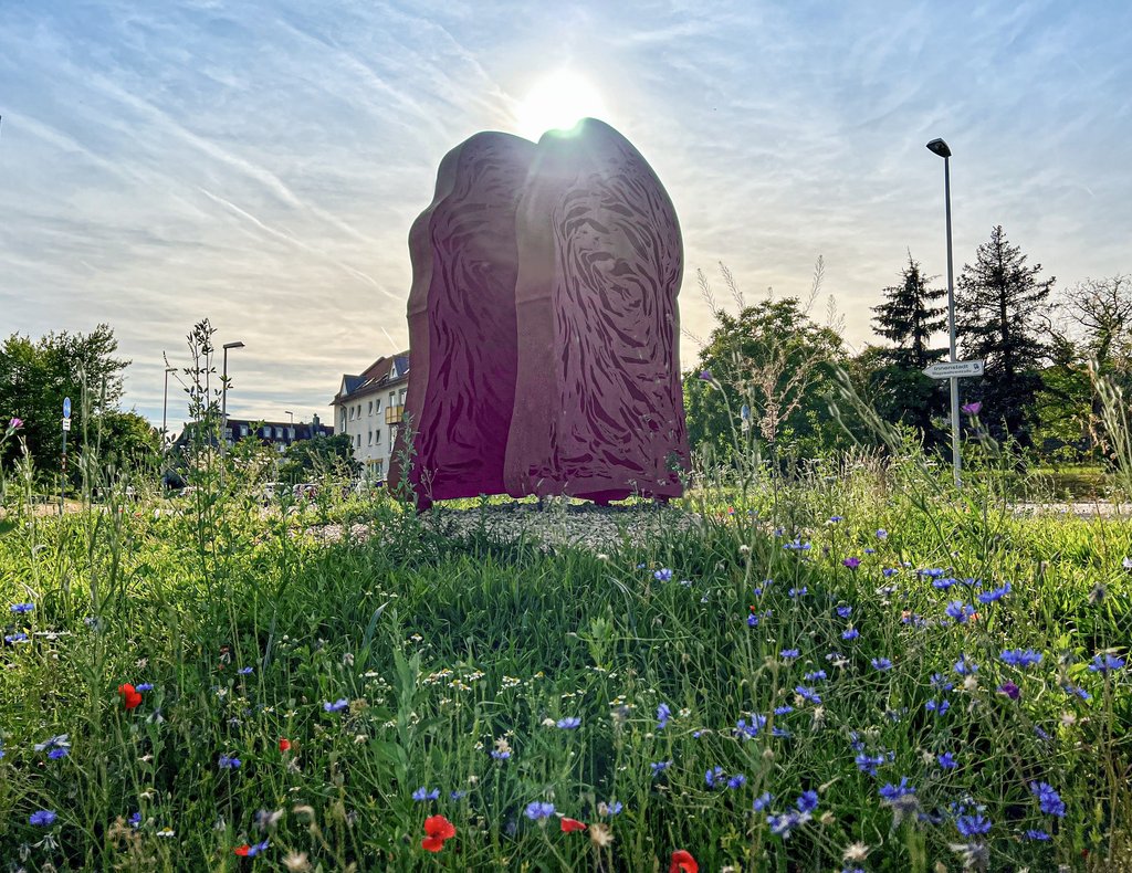 Auf der Darstellung ist eine Skulptur in Form von zwei versetzten Zwiebackscheiben auf einem Kreisel zu sehen.