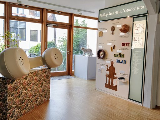 Das Foto zeigt das Foyer des Philipp-Reis-Hauses. In einem hellen Raum mit großen Fenstern steht links ein überdimensionaler Telefonhörer auf dem Empfangstisch. Rechts befindet sich eine Informationstafel zur Stadtgeschichte mit verschiedenen Objekten, Beschriftungen und dem Grundriss des Museums. Im Hintergrund ist durch die Glasfront ein begrünter Innenhof zu sehen.