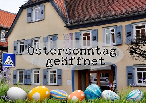 Vorderansicht eines zweistöckigen Hauses mit hellbeiger Fassade, blauen Fensterläden und einem roten Ziegeldach. Das Philipp-Reis-Haus. Im Vordergrund ist der Text ‚Ostersonntag geöffnet‘ zu sehen. Vor dem Haus sind bunt bemalte Ostereier in verschiedenen Größen und Mustern im Gras platziert.