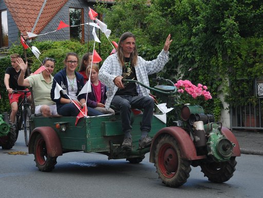 Das Bild zeigt eine fröhliche Szene bei einem vergangenen Blütenfest in Seulberg. Im Vordergrund fährt ein kleines, offenes Fahrzeug, das wie eine Mischung aus Traktor und Wagen aussieht. Am Steuer sitzt ein Mann, der lächelnd winkt. Auf der Ladefläche hinter ihm sitzen mehrere Kinder und Jugendliche, die ebenfalls in die Kamera schauen und teilweise winken. Hinter diesem Wagen fährt ein weiteres ähnliches Fahrzeug mit weiteren Personen.
