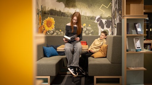Ein Mädchen und ein Junge befinden sich im Cocoon. Das Mädchen liest sitzend auf dem Tisch ein Buch. Der Junge sitzt auf der Bank während er an der Konsole spielt, diese steht auf dem Tisch.