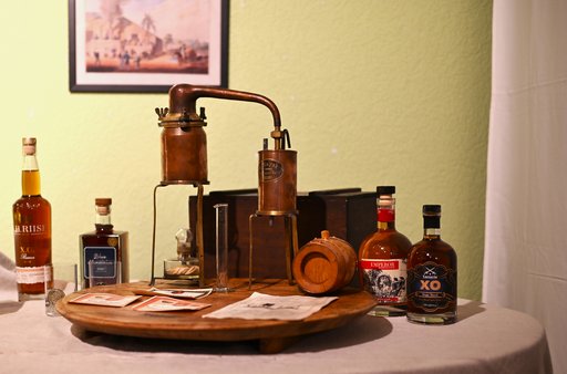 Zu sehen ist ein Tisch, mit Tischdecke auf dem ein Holztablett steht. Auf diesem Holztablett befindet sich eine kupferne, kleine Distille und ein kleines Rumfäßchen. Vor der kleinen Destille liegen verschiedene Etiketten und Papiere. Lunks und rechts neben dem Tablett stehen jeweils zwei verschiedene Rumflacshen in unterschiedlichen Größen. Hinter der Distille auf dem Tisch steht eine Holzkiste. Im Hintergrund ist ein Bild an der Wand zu erkennen.