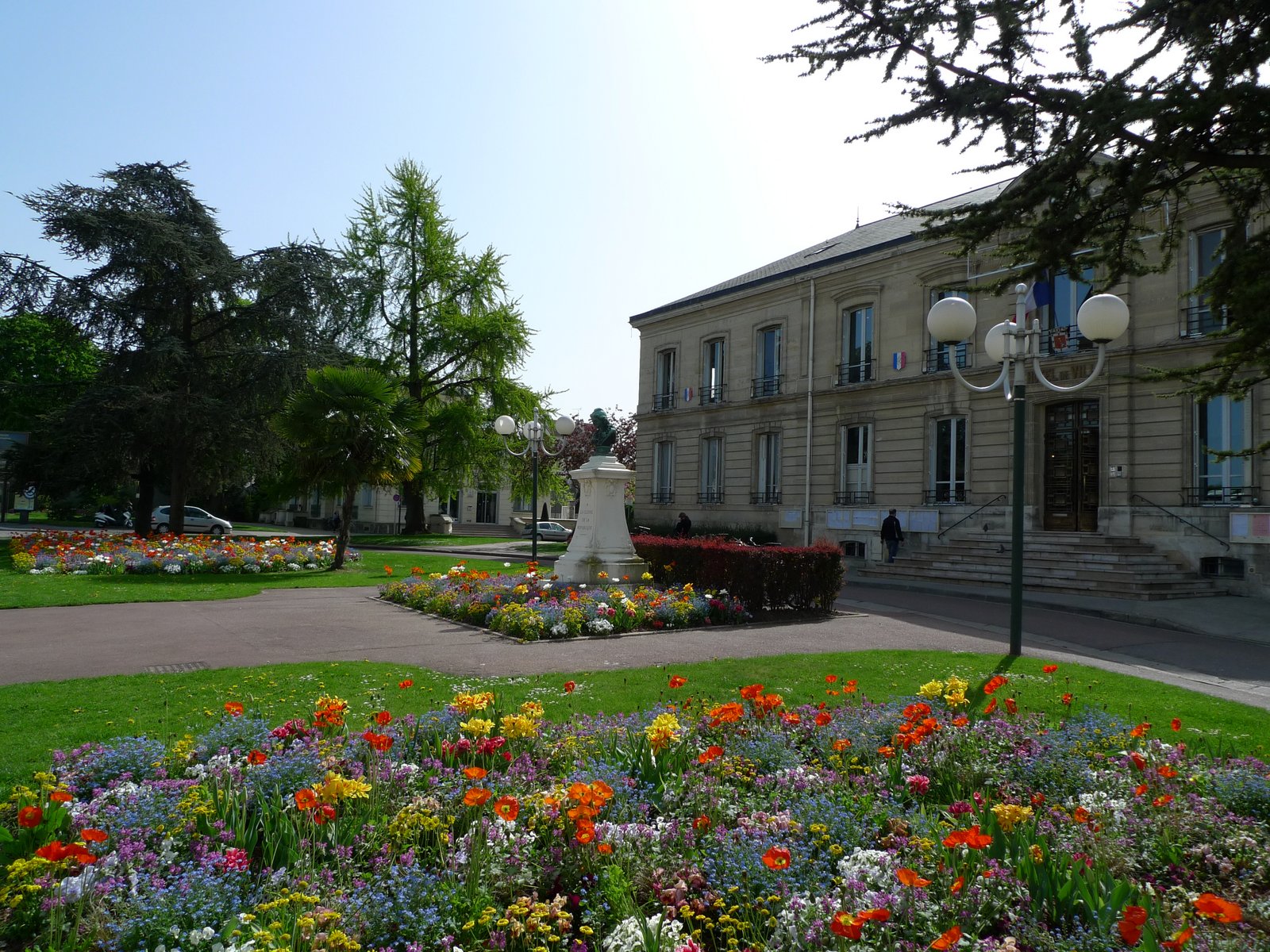 Auf der Darstellung ist das Rathaus von Houilles zu sehen, eingerahmt von Blumenbeeten.