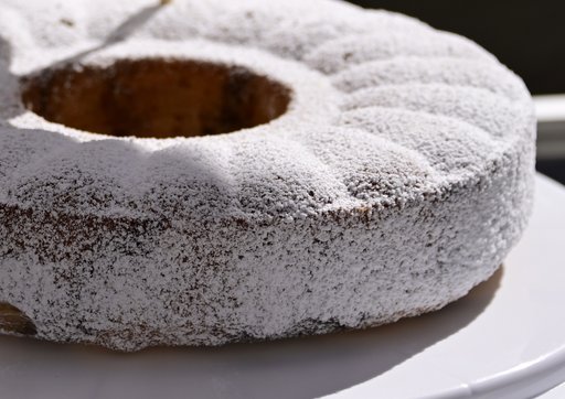 Das Bild zeigt einen Kuchen auf einem weißen Teller. Der Kuchen ist ringförmig mit einem Loch in der Mitte und vollständig mit Puderzucker bestäubt. Die Oberfläche des Kuchens ist gleichmäßig mit einer feinen Schicht Puderzucker bedeckt, der leicht glänzt.