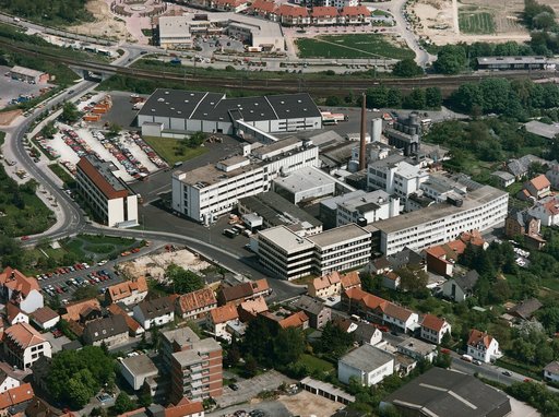 Luftaufnahme des Milupa-Firmengeländes in Friedrichsdorf, aufgenommen aus schräger Vogelperspektive an einem sonnigen Tag. Im Zentrum liegt ein dicht bebauter Fabrikkomplex aus mehrstöckigen, überwiegend weißen Gebäuden mit Flachdächern. Ein langgestreckter, leicht geschwungener Bau zieht sich am rechten Bildrand entlang. Dahinter ragen ein hoher, schmaler Backsteinschornstein und eine Gruppe grauer Silotürme auf. Im hinteren Bereich steht eine große, dunkel gedeckte Halle, vor der ein Lastwagen mit Milupa-Schriftzug parkt. Im Vordergrund stehen Wohnhäuser mit roten und grauen Dächern, in der Bahnstraße.