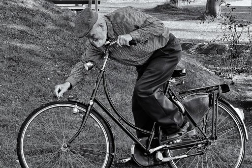 Senior , der zum Vorderrad seines Fahrrad greift im Vordergrund, Hafen/ Park im Hintergrund, schwarz-weiss Fotografie