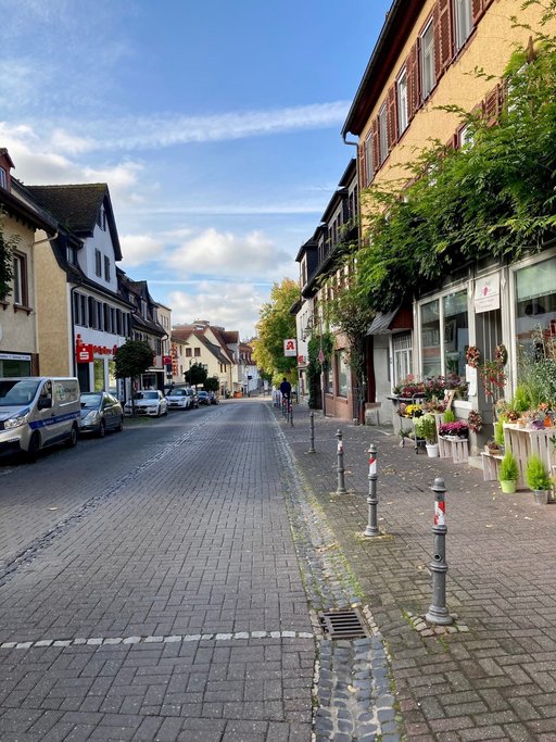 Das Bild zeigt einen Abschnitt der Hugenottenstraße mit parkenden Autos und Geschäften.
