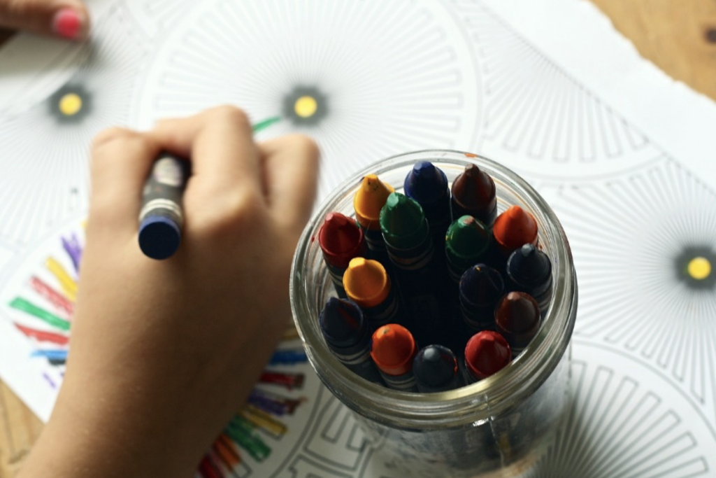 Auf der Darstellung ist eine Kinderhand zu erkennen, die mit Wachsmalstiften ein Bild ausmalt.