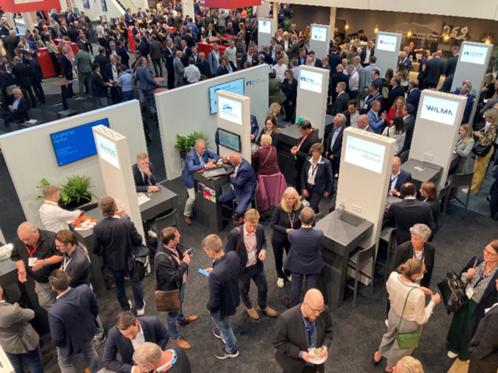 Viele Personen in Geschäftskleidung sind in Gesprächen auf einem Messegelände zu sehen.