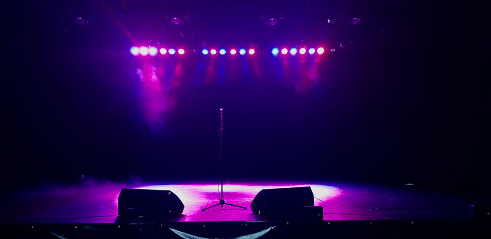 Auf der Darstellung ist die in pinkes und blaues Scheinwerferlicht getauchte Bühne im Forum Friedrichsdorf zu sehen, in der Mitte ein Mikrofonständer umrahmt von zwei Monitorboxen.
