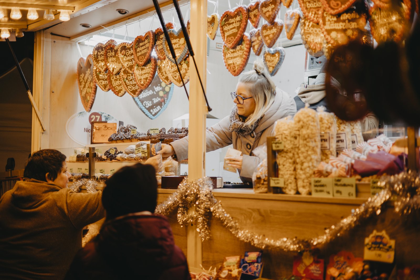 Weihnachtsmarkt 2024_Verkauf Das Bild zeigt einen Stand auf dem Friedrichsdorfer Weihnachtsmarkt. Im Vordergrund sind zwei Personen abgebildet die mit dem Rücken zum Betrachtenden gerade etwas an einem Süßwarenstand kaufen. Der Stand ist üppig mit Lebkuchenherzen und anderen Süßigkeiten dekoriert.