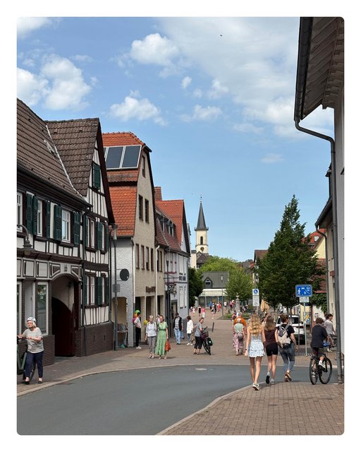 Das Bild ist mit dem Rücken zum Rathaus, neben dem Standesamt aufgenommen und zeigt den Blick auf den Landgrafenplatz und die Hugenottenstraße. Mehrere Menschen stehen und laufen auf dem Platz umher und im Hintergrund sieht man den Kirchturm.