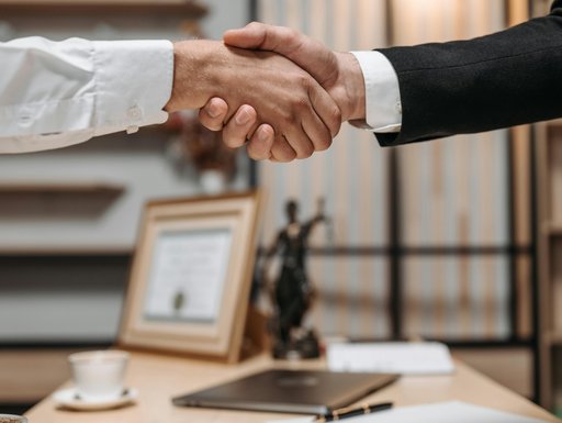 Zwei Personen reichen sich in einem Büro die Hand. Eine Person trägt ein weißes Hemd, die andere einen schwarzen Anzug. Auf dem Schreibtisch im Hintergrund stehen eine Tasse, ein Laptop, Schreibutensilien sowie ein Bilderrahmen. Außerdem ist eine Justitia-Figur sichtbar, was auf ein juristisches Umfeld hinweist.