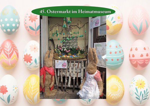 Plakat zum „45. Ostermarkt im Heimatmuseum“. In einem dekorierten Schaufenster stehen zwei Strohhasenfiguren – links mit rotem Tuch, rechts mit Kleid und Schürze – vor einem kleinen Zaun aus Birkenzweigen. Darüber hängen grün bemalte Ostereier an Ästen. In der Mitte befindet sich ein Osterarrangement mit Frühlingsblumen und bunten Eiern. Der Hintergrund ist mit großen, pastellfarbenen, verzierten Ostereiern gestaltet.