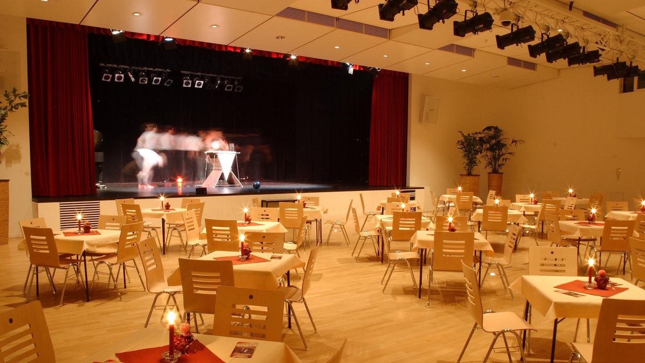 Auf der Darstellung ist der große Saal mit stilvoll eingedeckten Tischen zu sehen, auf der Bühne ein Künstler der Varieté-Show.