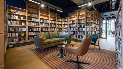 Eine Sitzgruppe mit einer Couch, zwei Sesseln, zwei Tischen und einer Pflanze, im Hintergrund eine Regalwand mit Büchern.